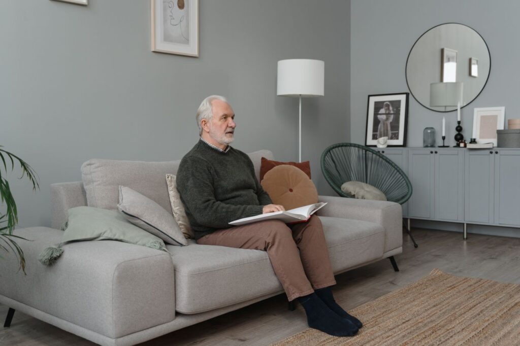 Elderly man reading a Braille book while sitting on a comfortable sofa in a modern living room.